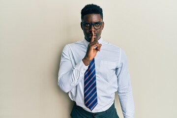 Fototapeta premium Handsome black man wearing glasses business shirt and tie asking to be quiet with finger on lips. silence and secret concept.