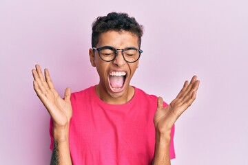 Young handsome african american man wearing glasses over pink background celebrating mad and crazy for success with arms raised and closed eyes screaming excited. winner concept