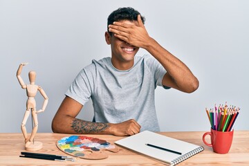 Young handsome african american man painter sitting palette and art manikin smiling and laughing with hand on face covering eyes for surprise. blind concept.