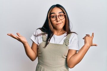 Young latin woman wearing casual clothes and glasses clueless and confused expression with arms and...