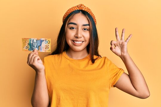 Young latin woman holding 50 australian dollar banknote doing ok sign with fingers, smiling friendly gesturing excellent symbol