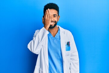 Handsome hispanic man with beard wearing doctor uniform covering one eye with hand, confident smile on face and surprise emotion.