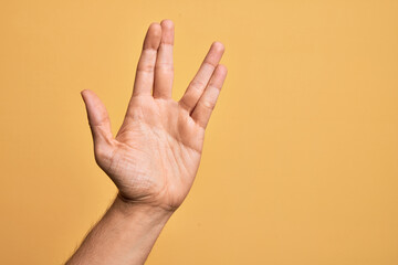 Hand of caucasian young man showing fingers over isolated yellow background greeting doing Vulcan salute, showing hand palm and fingers, freak culture