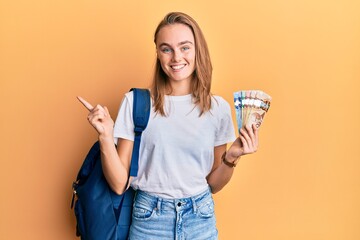 Beautiful blonde woman wearing student backpack and holding canadian dollars smiling happy pointing...