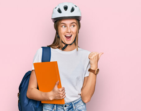 Beautiful young blonde woman wearing backpack and bike helmet pointing thumb up to the side smiling happy with open mouth
