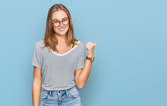 Beautiful young blonde woman wearing casual clothes and glasses smiling with happy face looking and pointing to the side with thumb up.