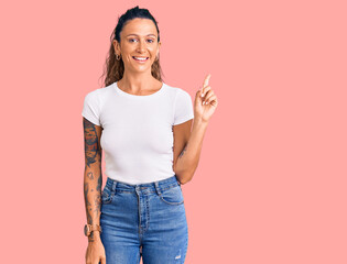 Young hispanic woman with tattoo wearing casual white tshirt with a big smile on face, pointing with hand finger to the side looking at the camera. - Powered by Adobe