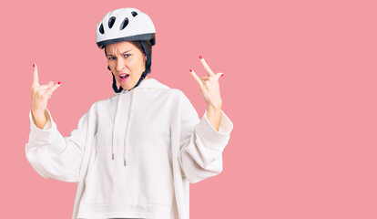 Beautiful brunette young woman wearing bike helmet and sporty clothes shouting with crazy expression doing rock symbol with hands up. music star. heavy concept.