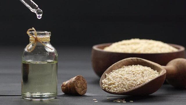 Glass bottle of sesame oil and raw sesame seeds in wooden shovel on black wooden table. Oil dripping