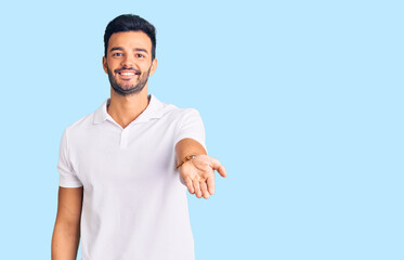 Young handsome hispanic man wearing casual clothes smiling cheerful offering palm hand giving assistance and acceptance.