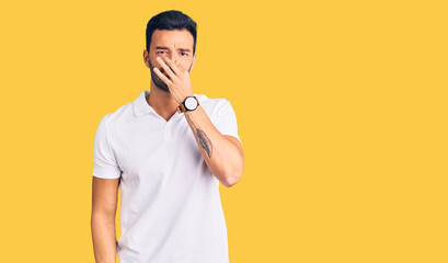 Young handsome hispanic man wearing casual clothes smelling something stinky and disgusting,...