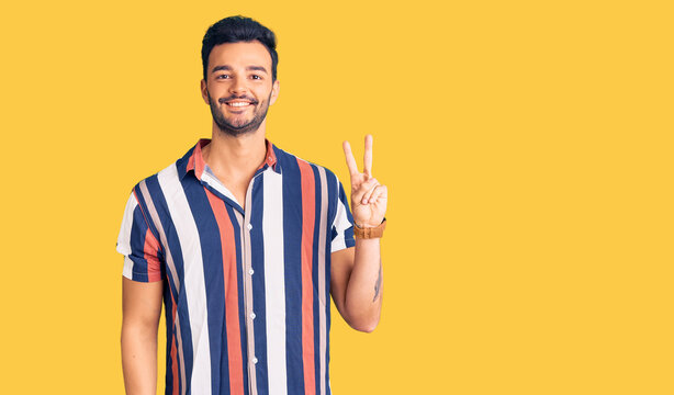 Young handsome hispanic man wearing casual  summer shirt smiling with happy face winking at the camera doing victory sign. number two.