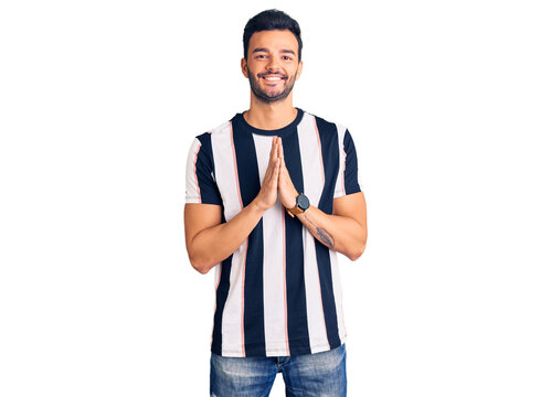 Young handsome hispanic man wearing striped tshirt praying with hands together asking for forgiveness smiling confident.