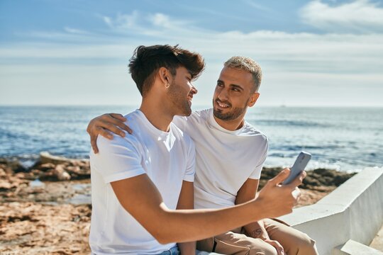 Young gay couple smiling happy using smartphone at the beach.