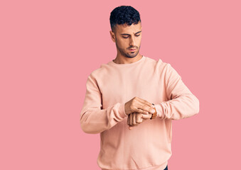 Young hispanic man wearing casual clothes checking the time on wrist watch, relaxed and confident
