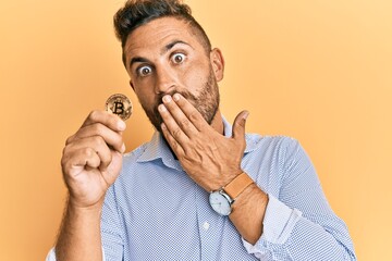 Handsome man with beard holding virtual currency bitcoin covering mouth with hand, shocked and...