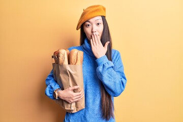 Young beautiful chinese woman wearing french beret holding paper bag with bread covering mouth with hand, shocked and afraid for mistake. surprised expression