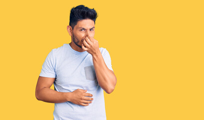 Handsome latin american young man wearing casual clothes smelling something stinky and disgusting,...