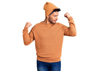 Naklejka premium Handsome latin american young man wearing winter sweater and wool hat showing arms muscles smiling proud. fitness concept.