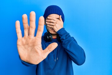 Naklejka premium Hispanic young man wearing sweatshirt and headphones covering eyes with hands and doing stop gesture with sad and fear expression. embarrassed and negative concept.
