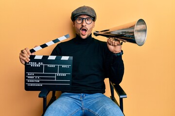 Handsome man with tattoos holding video film clapboard and louder afraid and shocked with surprise...