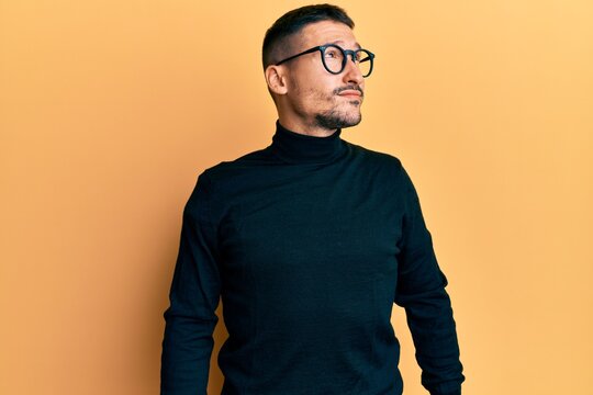 Handsome man with tattoos wearing turtleneck sweater and glasses smiling looking to the side and staring away thinking.