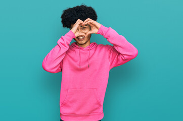 Young african american man with afro hair wearing casual pink sweatshirt doing heart shape with hand and fingers smiling looking through sign