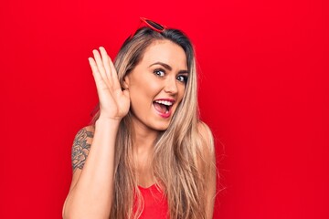 Young beautiful blonde woman wearing red t-shirt and sunglasses over isolated background smiling with hand over ear listening and hearing to rumor or gossip. Deafness concept.