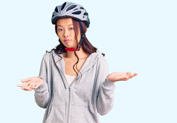 Young beautiful chinese girl wearing bike helmet clueless and confused expression with arms and hands raised. doubt concept.