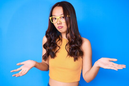 Young beautiful chinese girl wearing casual clothes and glasses clueless and confused expression with arms and hands raised. doubt concept.