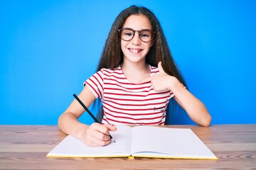 Cute hispanic child girl sitting on the table writing book pointing finger to one self smiling happy and proud