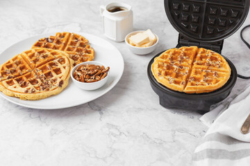 Homemade Pecan Waffles on a White Plate; One Still in Waffle Iron; White Countertop; Butter and Maple Syrup in Background