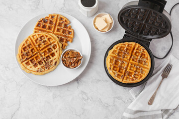 Homemade Pecan Waffles on a White Plate; One Still in Waffle Iron; White Countertop; Butter and Maple Syrup in Background