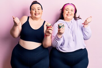 Young plus size twins wearing sportswear holding cupcake screaming proud, celebrating victory and...