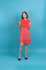 full-length smiling happy young asian woman in red dress holding big red paper heart isolated on blue background.