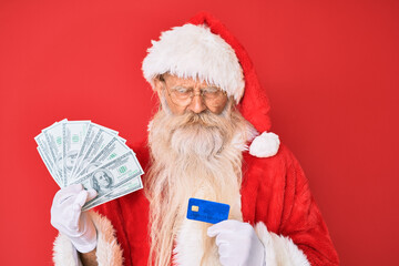 Old senior man wearing santa claus costume holding dollars and credit card skeptic and nervous, frowning upset because of problem. negative person.