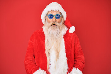 Old senior man wearing santa claus costume and sunglasses scared and amazed with open mouth for surprise, disbelief face