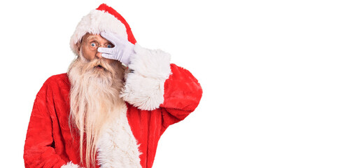 Old senior man with grey hair and long beard wearing traditional santa claus costume peeking in shock covering face and eyes with hand, looking through fingers with embarrassed expression.