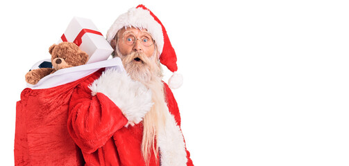 Old senior man with grey hair and long beard wearing santa claus costume holding bag with presents...