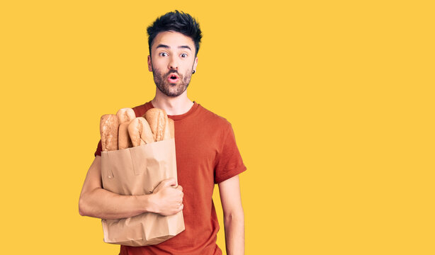Young hispanic man holding paper bag with bread scared and amazed with open mouth for surprise, disbelief face