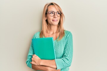 Beautiful blonde woman holding book wearing glasses smiling looking to the side and staring away thinking.