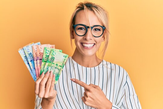 Beautiful blonde woman holding south african rand banknotes smiling happy pointing with hand and finger