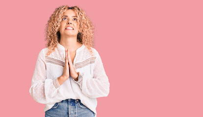 Young blonde woman with curly hair wearing elegant summer shirt begging and praying with hands...