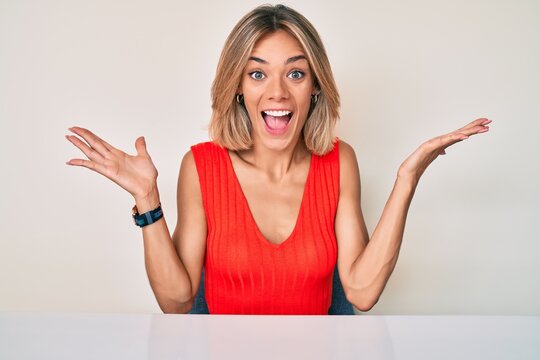 Beautiful caucasian woman wearing casual clothes sitting on the table celebrating victory with happy smile and winner expression with raised hands