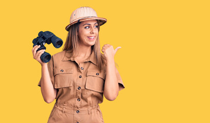 Young beautiful woman wearing explorer hat holding binoculars pointing thumb up to the side smiling happy with open mouth