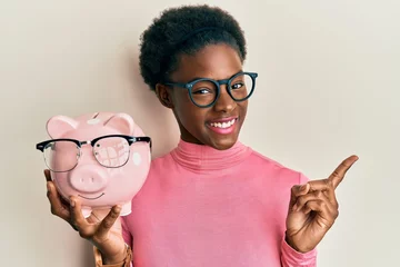 Fotobehang Young african american girl holding piggy bank with glasses smiling happy pointing with hand and finger to the side © Krakenimages.com