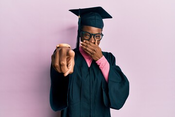 Young african american girl wearing graduation cap and ceremony robe laughing at you, pointing...