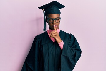 Young african american girl wearing graduation cap and ceremony robe asking to be quiet with finger on lips. silence and secret concept.