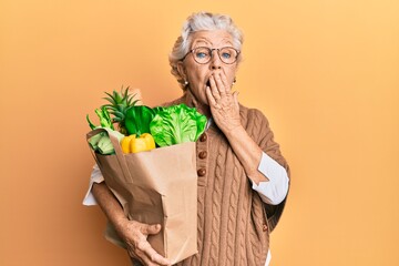 Senior grey-haired woman holding paper bag with bread and groceries covering mouth with hand,...