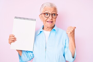 Senior beautiful woman with blue eyes and grey hair holding notebook pointing thumb up to the side smiling happy with open mouth